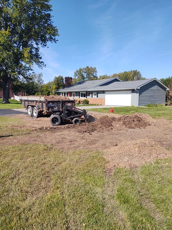A utility trailer with mulch sits in a yard next to a house under a blue sky.