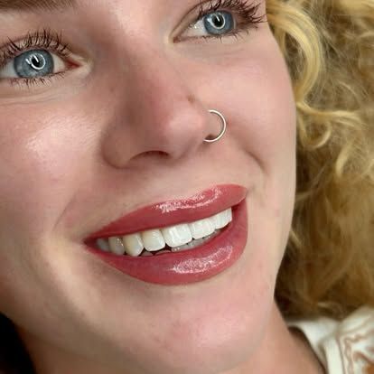 Close-up of a smiling person with curly blond hair, blue eyes, glossy pink lips, and a nose ring