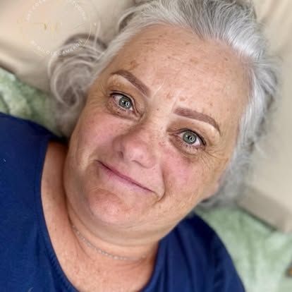 Smiling person with white hair in a blue shirt, lying on a bed or couch.
