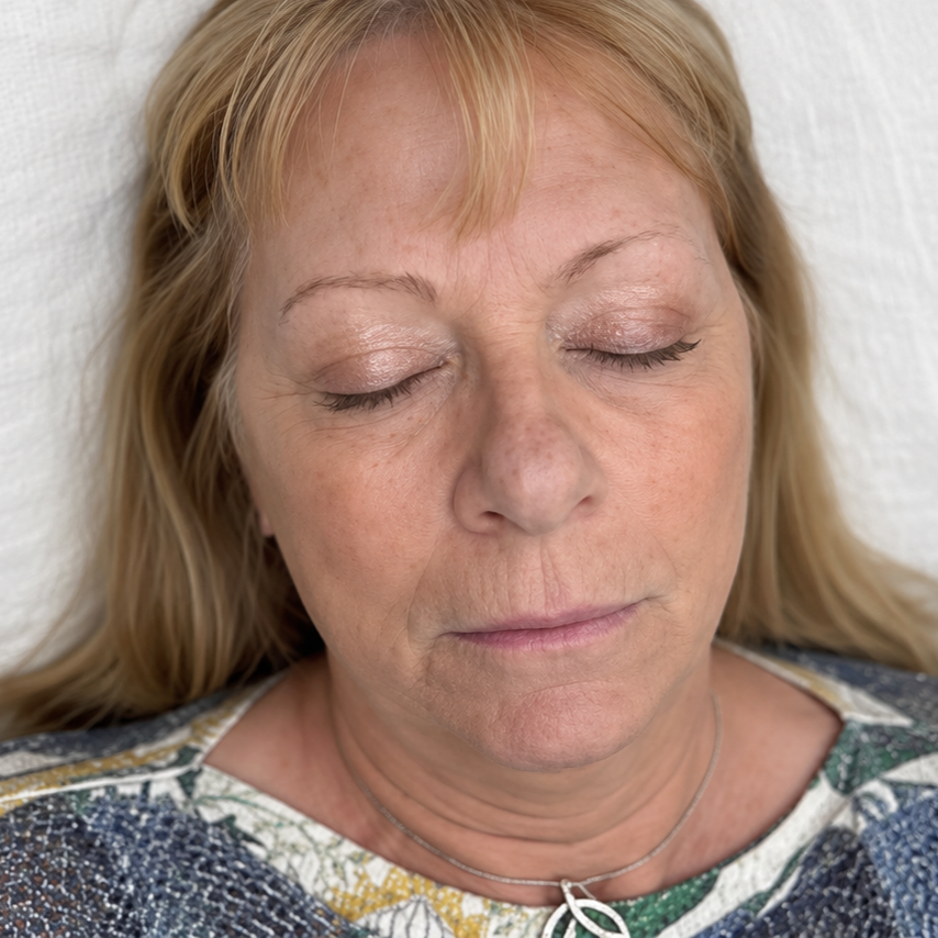 Woman lying on a bed with eyes closed, wearing a patterned top and necklace.