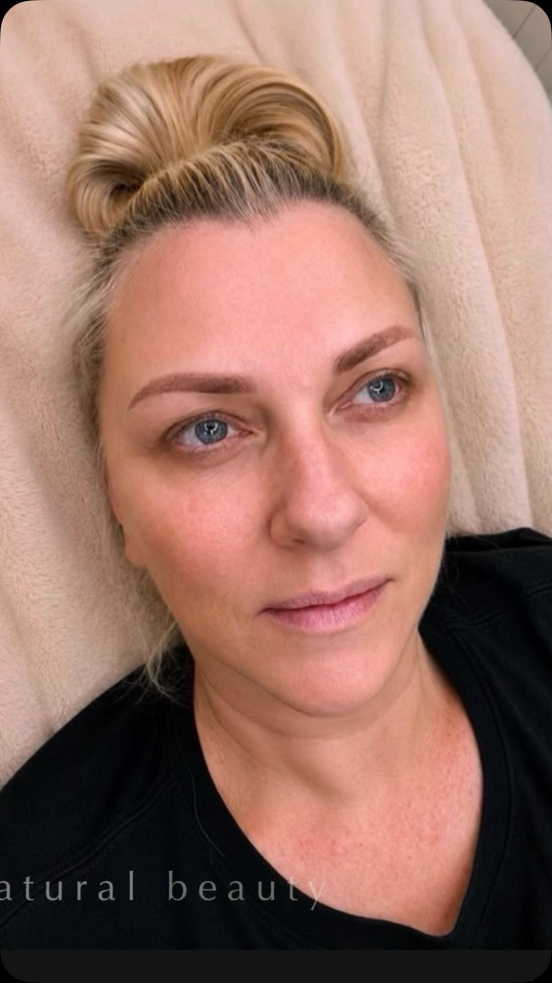 Close-up portrait of a woman with blonde hair in a bun, lying on a beige cushion, wearing a black top.