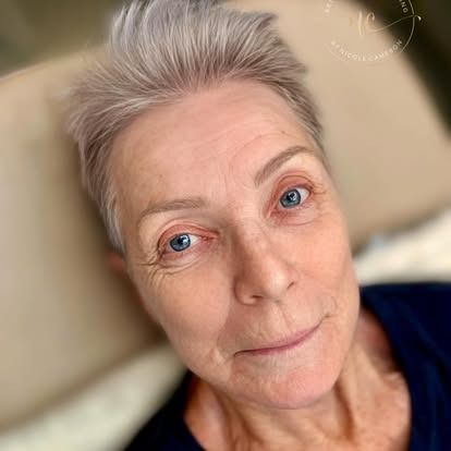 Close-up portrait of a person with short gray hair and blue eyes against a softly blurred indoor background
