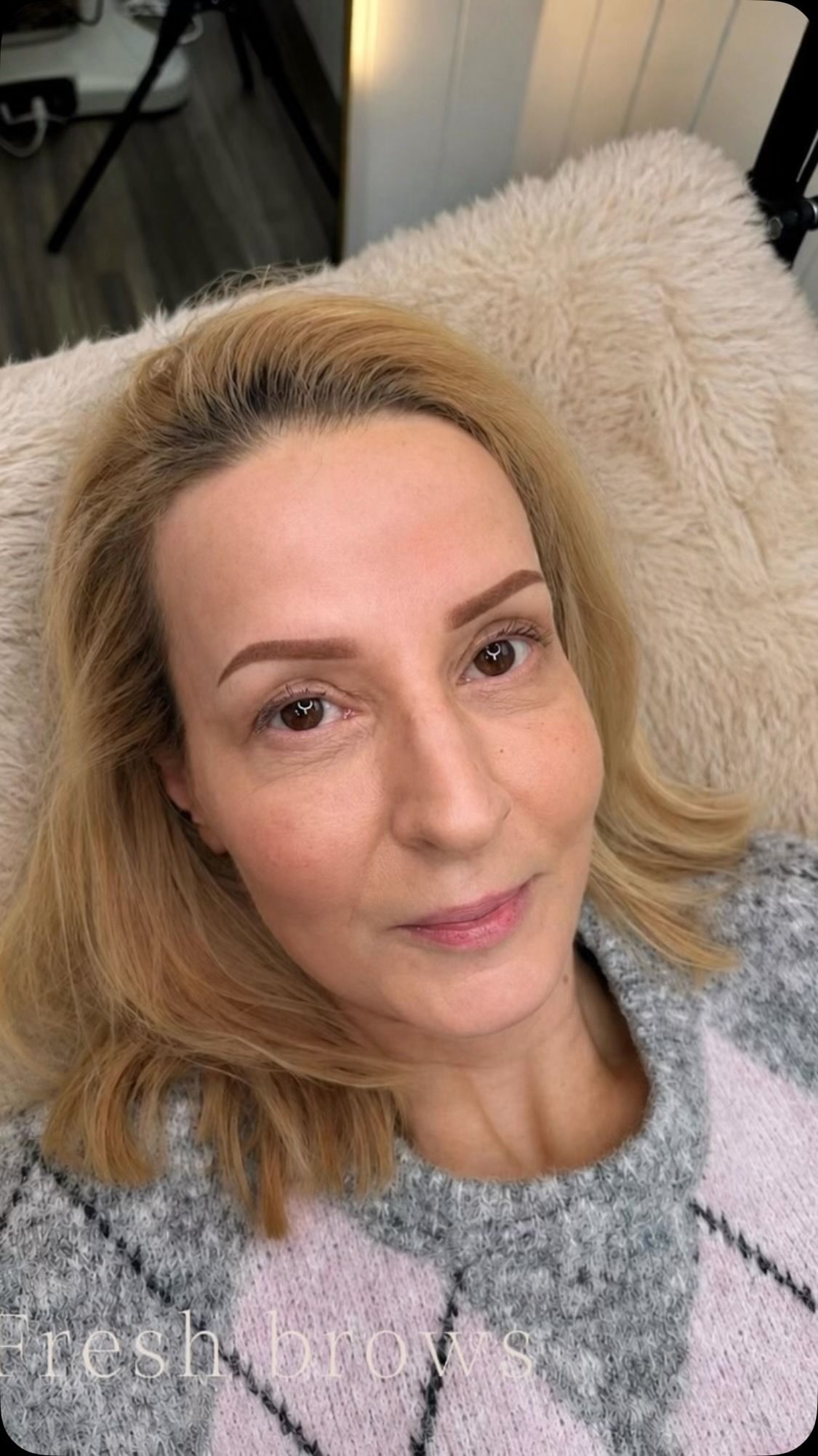 Close-up selfie of a person with blond hair resting on a fluffy beige chair, wearing a gray sweater, looking at the camera