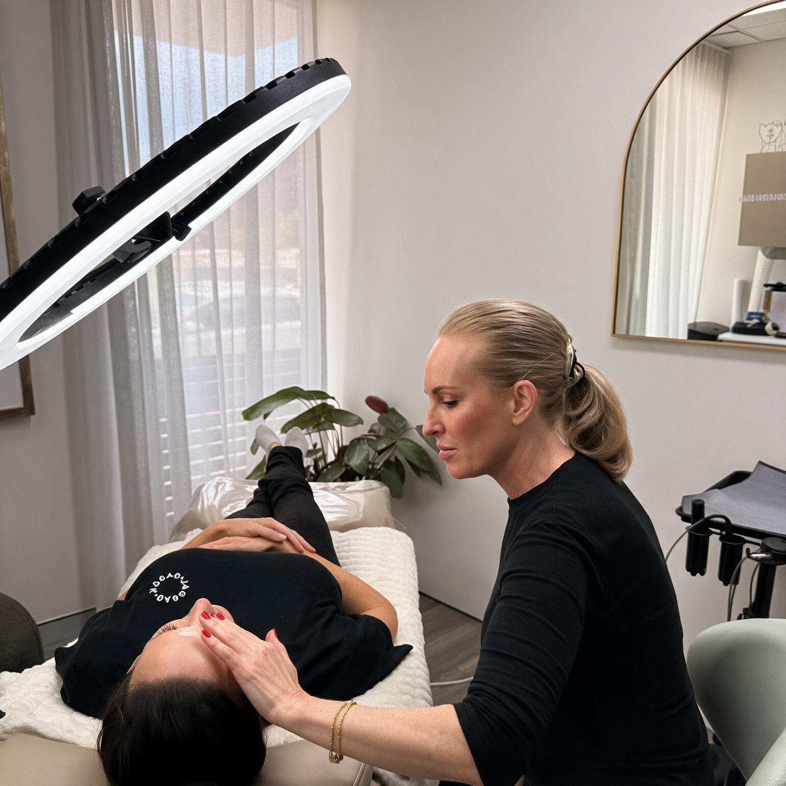 Woman performing a facial treatment on a client in a spa room under a bright ring light