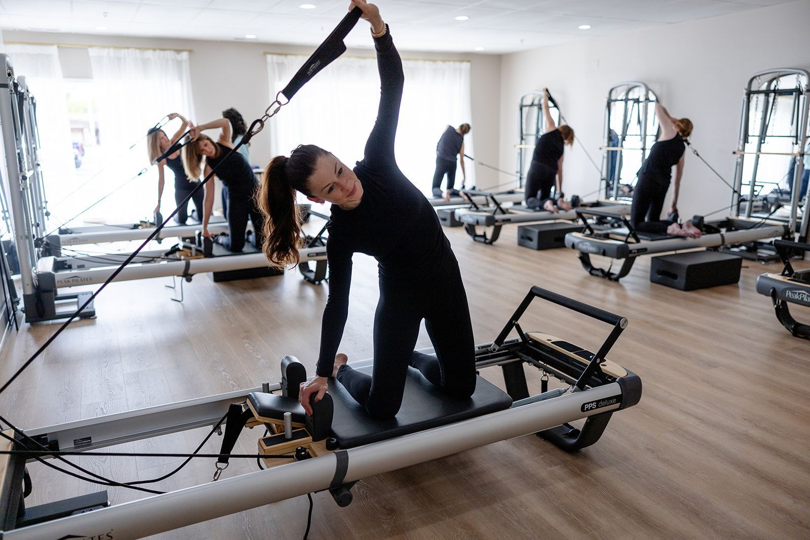 A Pilates class in a bright studio. People in black workout clothes stretch on reformer machines.