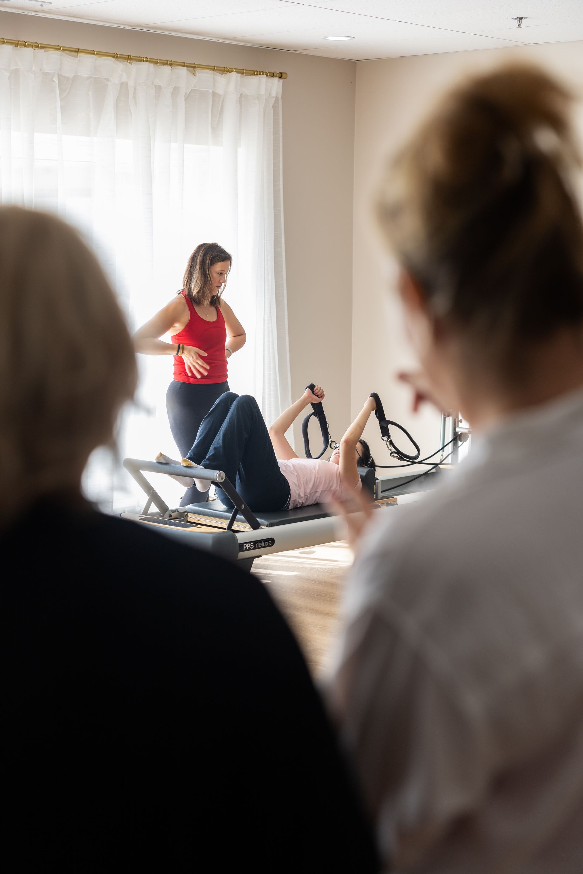 Pilates instructor guides client on machine, two others watch in bright room.