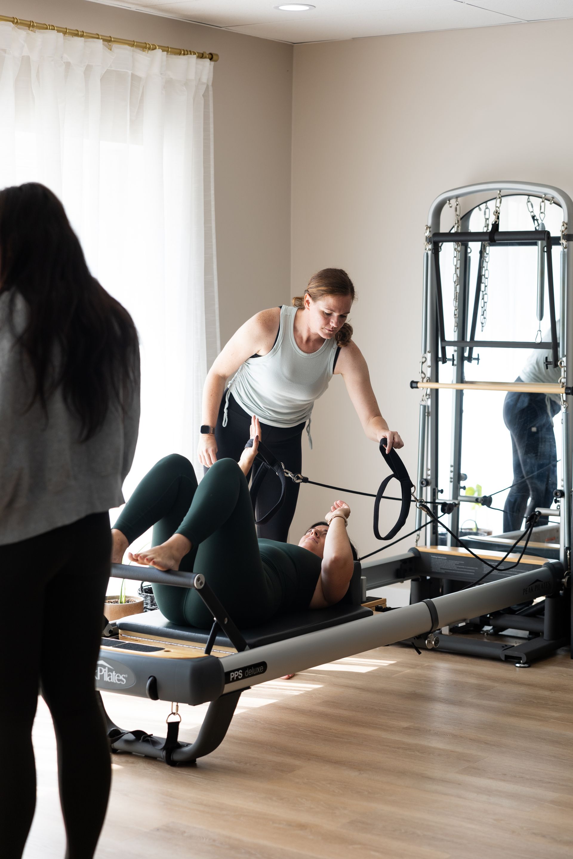 Pilates session on a Pilates reformer in a bright studio, with a trainer assisting a client.