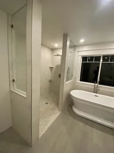 A modern bathroom with a white freestanding tub, a walk-in tiled shower with glass walls, and a window with a dark view.