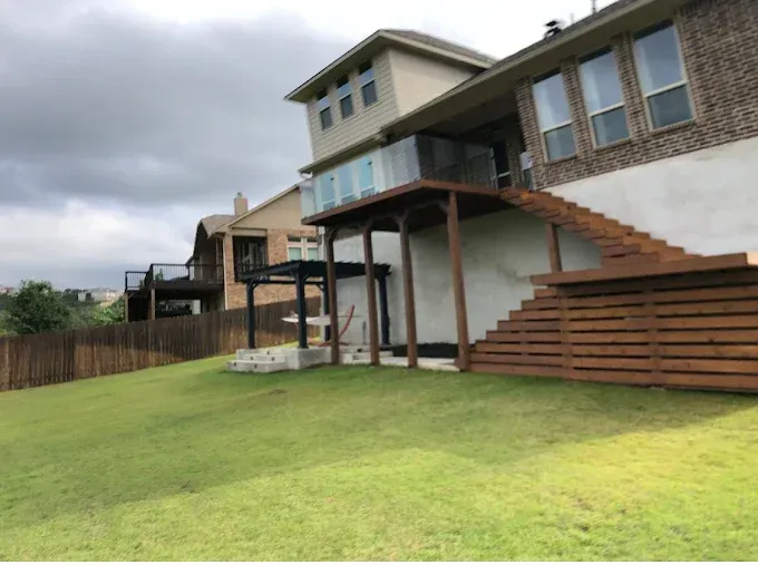 Rear view of a house featuring a wooden deck with a glass railing and wide stairs descending to a grassy backyard.