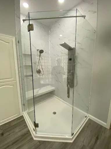 A glass neo-angle shower enclosure featuring white marble-patterned walls, a corner bench, and a brushed metal shower panel.