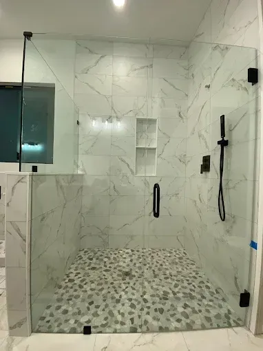 A glass-enclosed shower with white marble-patterned tile walls, a pebble-stone floor, black fixtures, and a dual-niche.