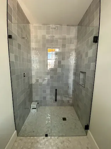 A walk-in shower with gray tiled walls, a glass door, a dark handle, a small niche, and a light-colored tiled floor.