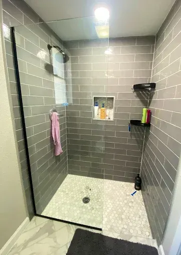 A modern walk-in shower with gray subway tile walls, a white pebble floor, a glass panel, and a wall-mounted niche.
