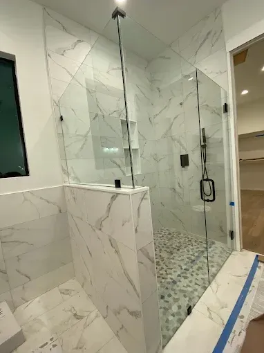 A modern walk-in shower with white marble-patterned tile walls, a glass enclosure, and a pebble-stone floor.