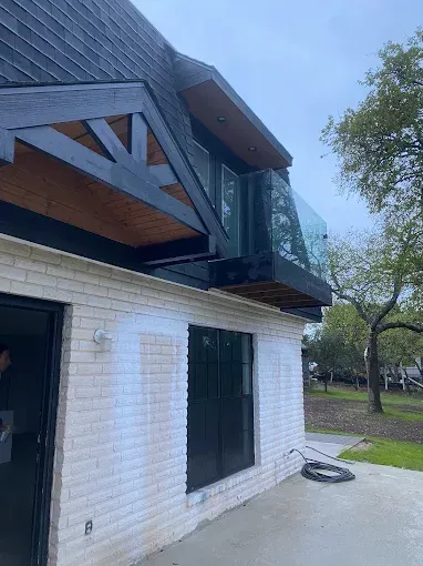 A modern exterior renovation featuring a white-painted brick wall, dark wood gable, and a glass-paneled balcony.