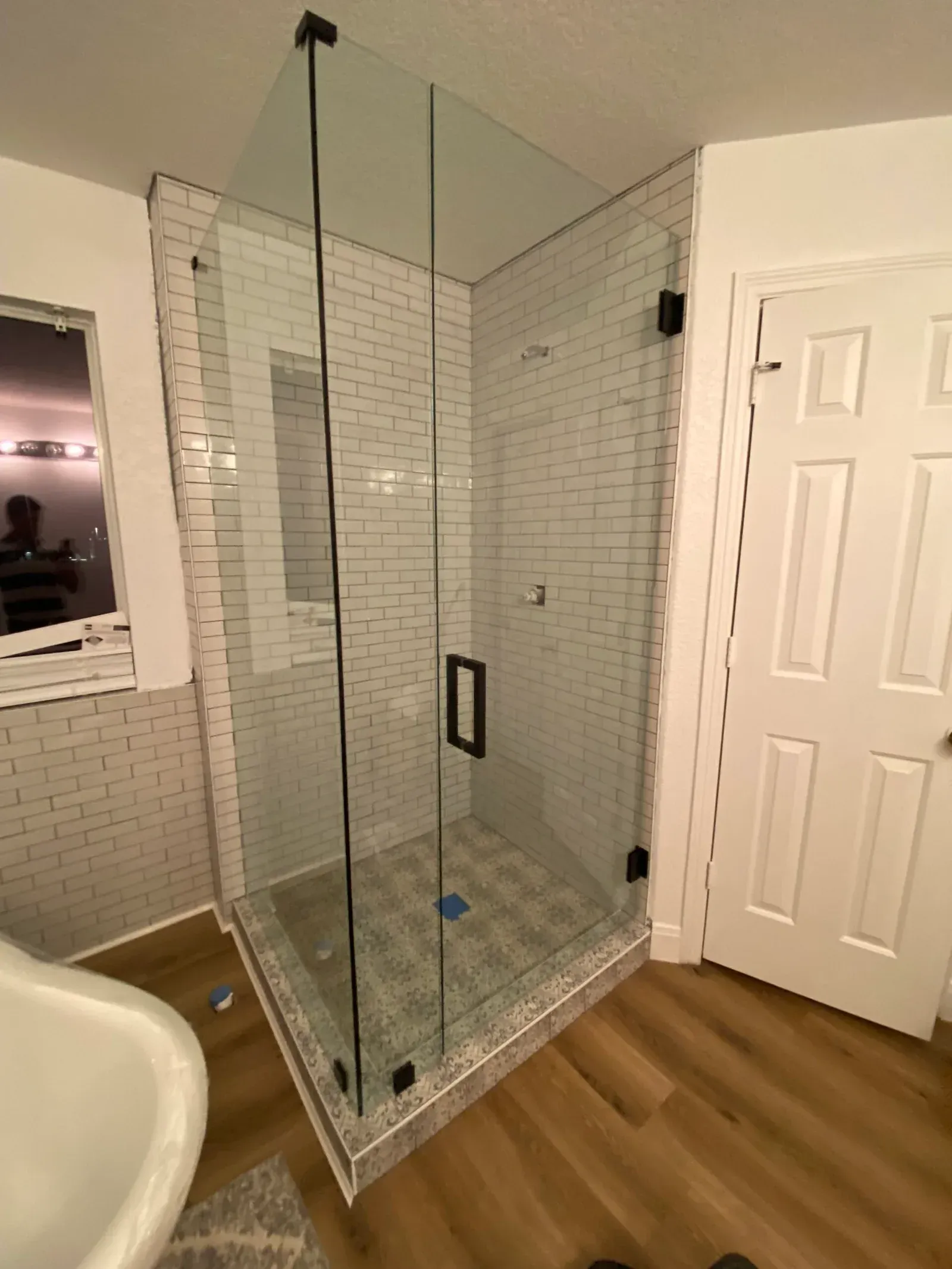A corner glass shower stall with black hardware, set against white subway tiles and wood-look flooring in a bathroom.