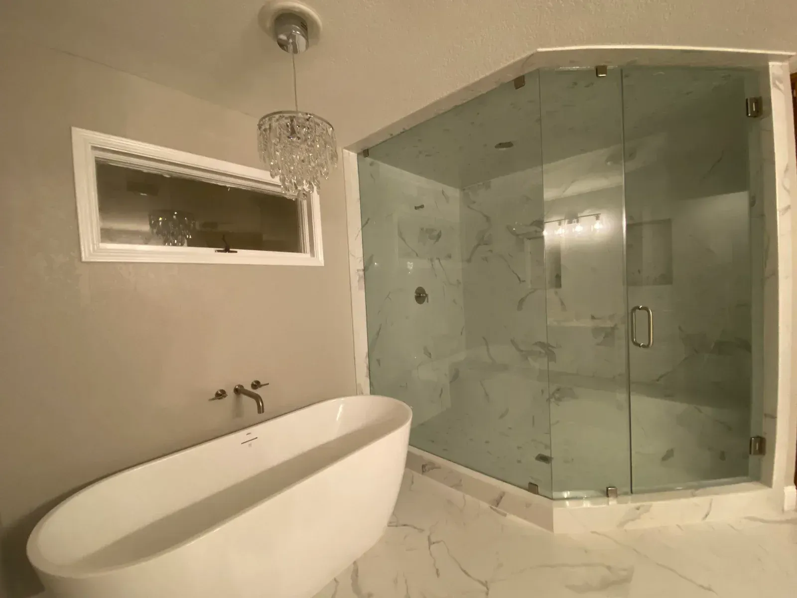 A modern bathroom featuring a white freestanding bathtub, a glass-enclosed shower, and a crystal chandelier.