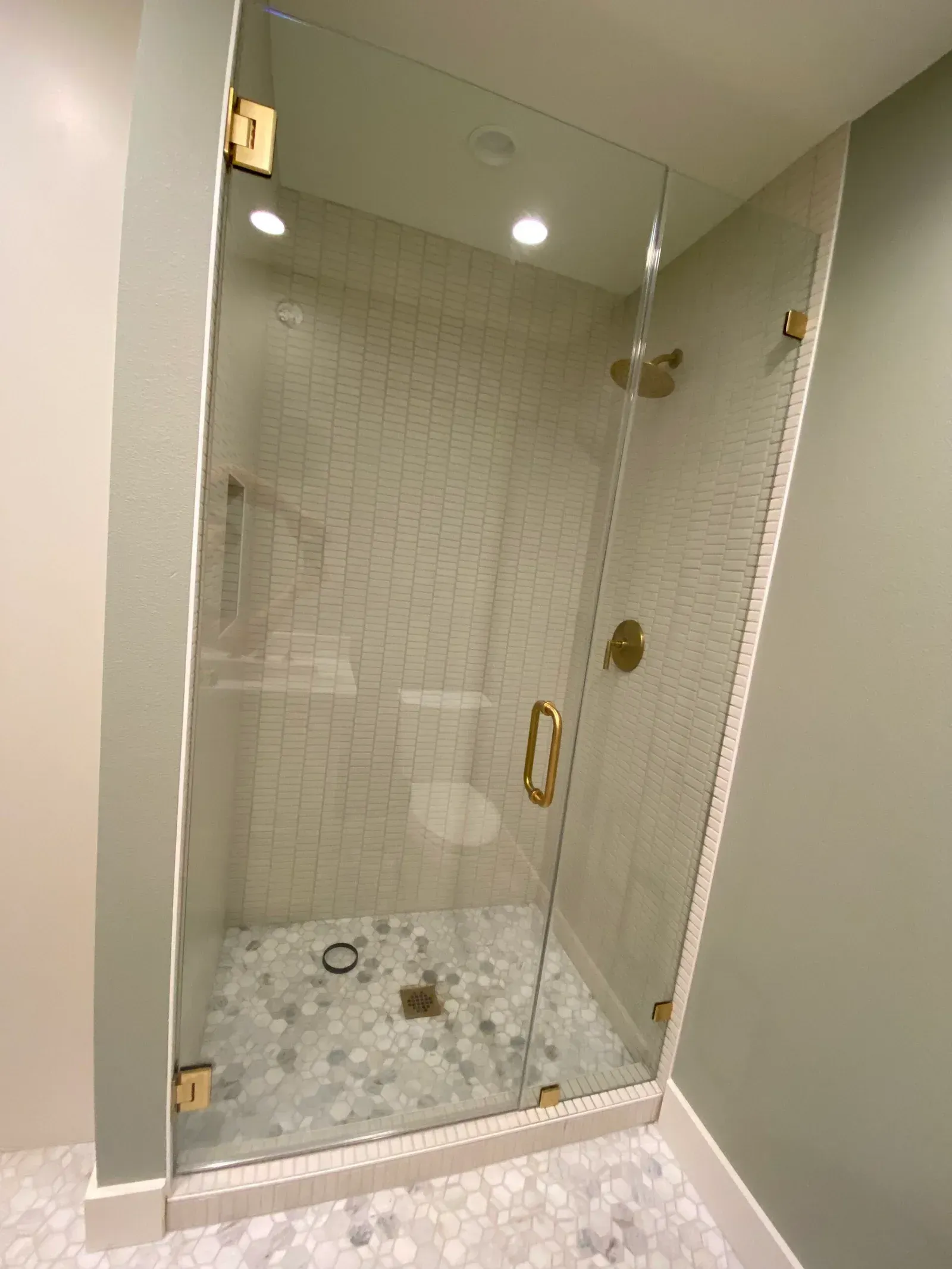 A glass shower enclosure with gold fixtures and a tiled pebble floor, set against sage green walls.