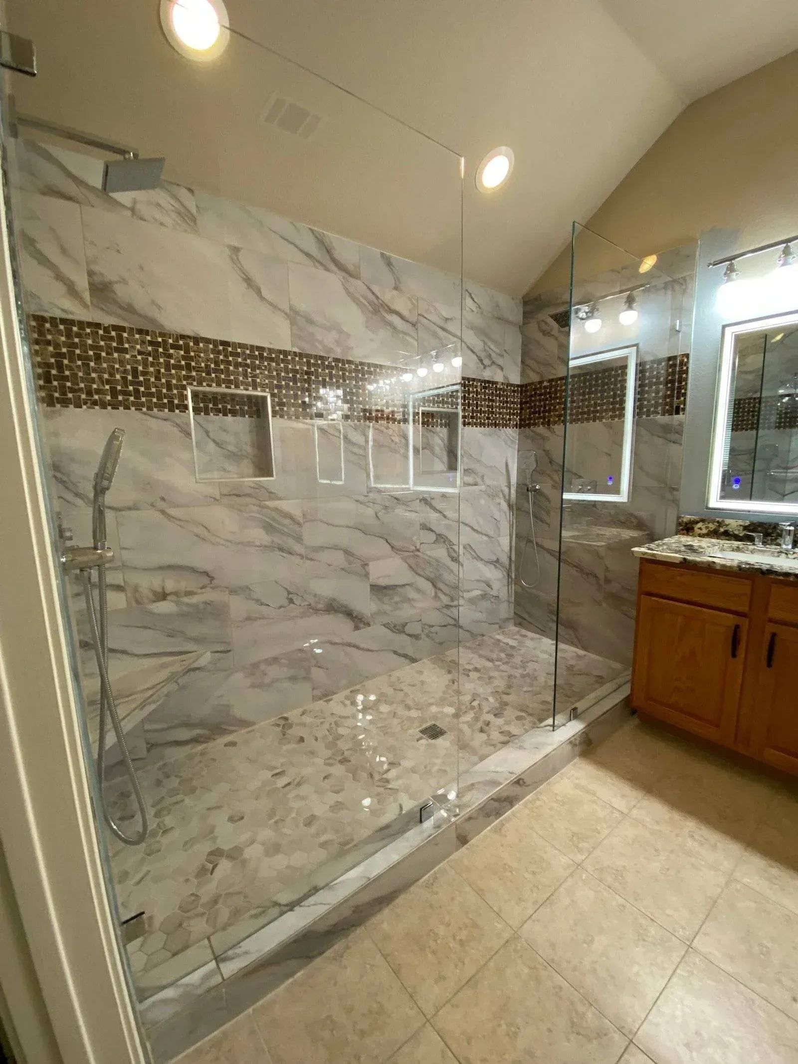 A modern walk-in shower with marbled walls, a glass enclosure, a mosaic accent tile strip, and a wood vanity with a mirror.