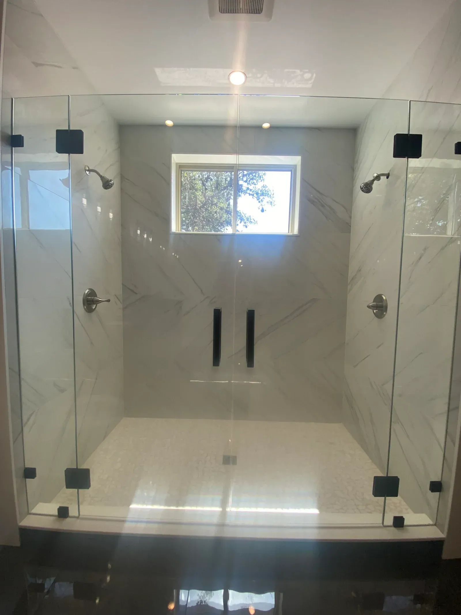 Walk-in shower with marble-style walls, glass doors, black hardware, and two showerheads in a white-tiled space.