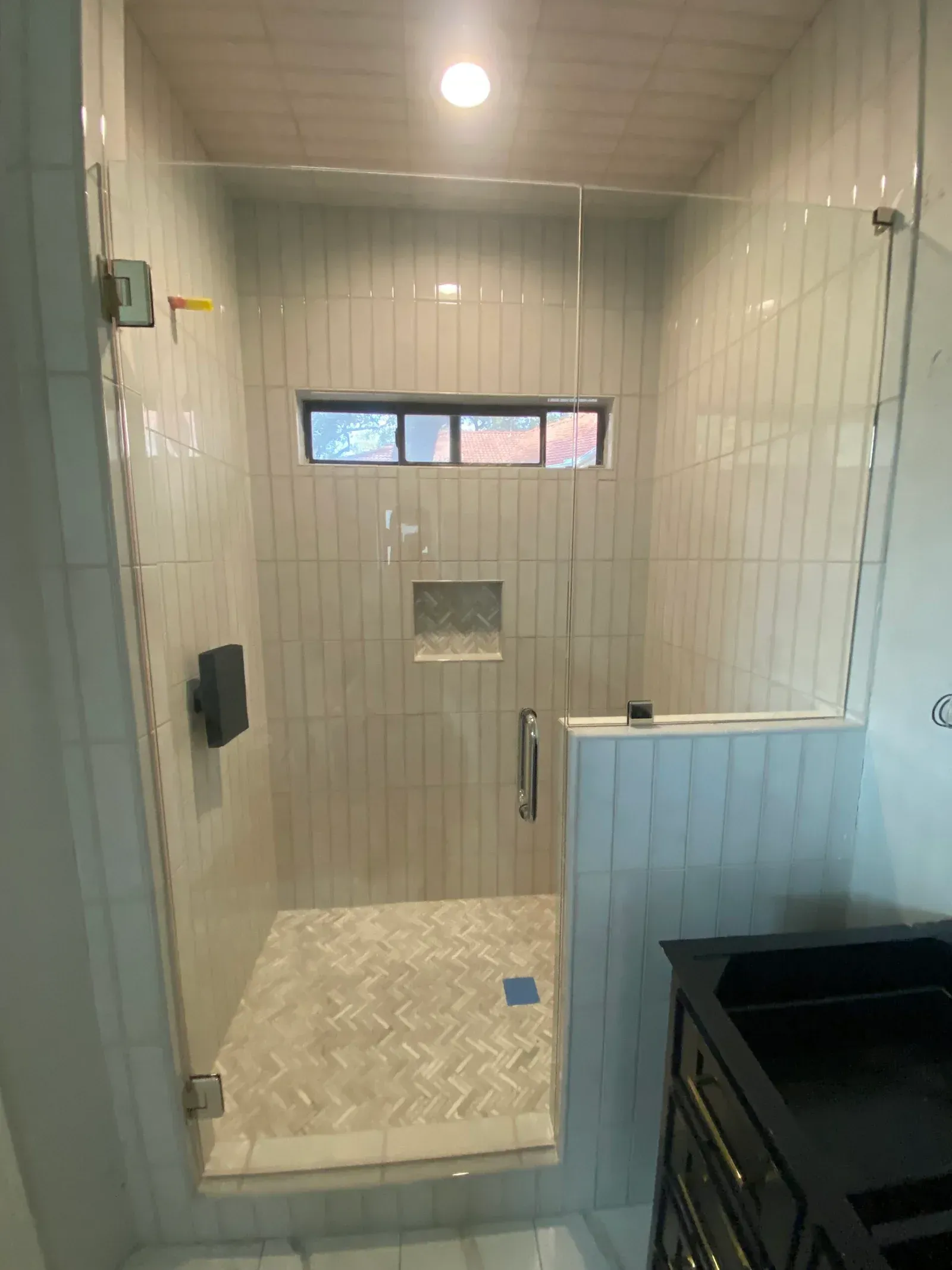 A walk-in shower with vertical white subway tiles, a small window, a niche, and glass doors beside a dark vanity.
