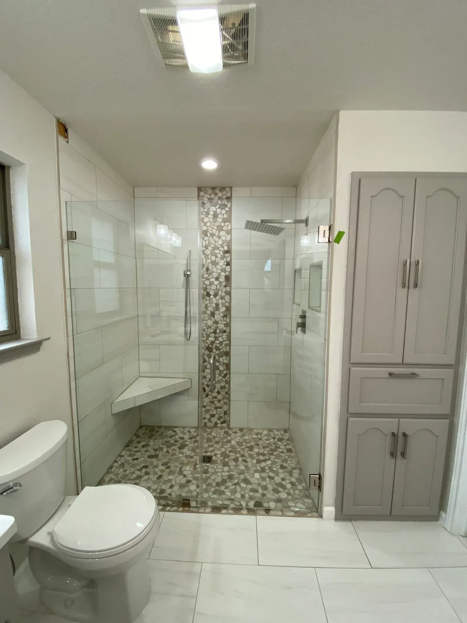 A bathroom featuring a walk-in shower with a mosaic tile floor, glass walls, and a matching tiled column, next to a cabinet.