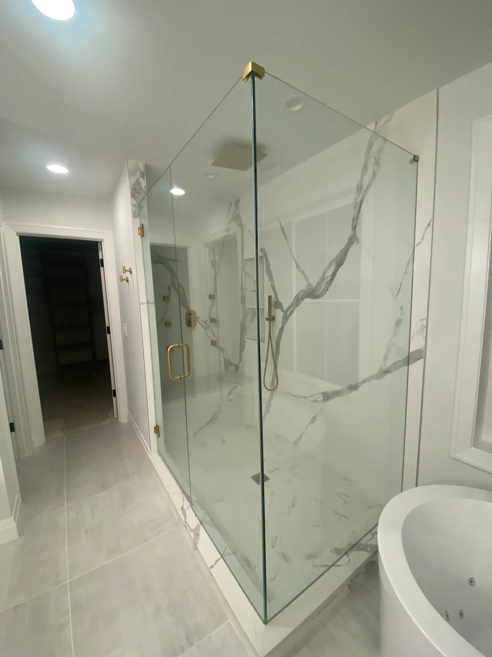 A modern glass shower enclosure with gold hardware, set against white marble-patterned walls in a bright bathroom.