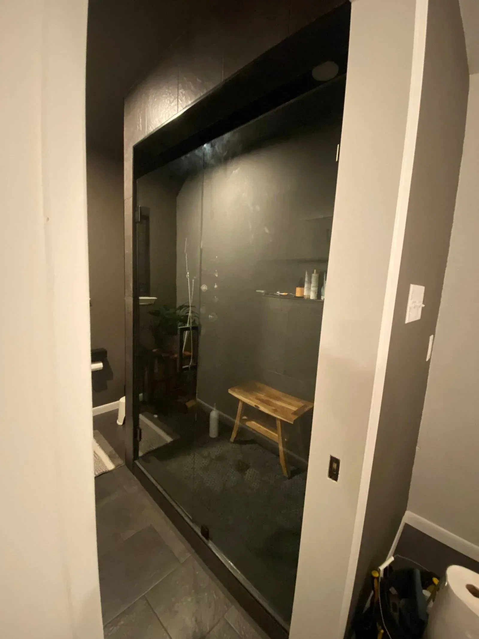 A modern walk-in shower with dark grey tiles, a glass enclosure, and a small wooden bench inside.