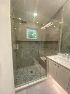 A modern bathroom featuring a glass-enclosed shower with pebble-textured flooring, gray wall tiles, and a white vanity.