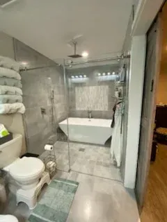 A modern bathroom with a white freestanding tub, glass-enclosed shower, light gray tile, and a white toilet.