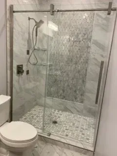 A modern bathroom with a glass sliding shower door featuring a hand-held showerhead, mosaic tile floor, and white toilet.