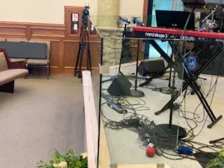 A Nord Stage 3 keyboard on a stand behind a glass partition, with many cables and equipment on the floor of a church stage.
