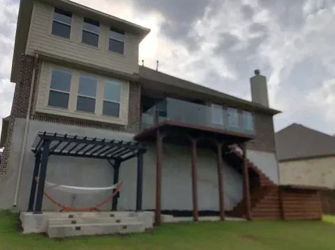 Back of a two-story house with a backyard deck, glass railing, wood stairs, and a pergola with a hammock on a patio.