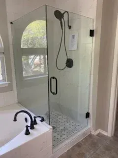 A modern walk-in shower with glass doors, black fixtures, and mosaic floor tile next to a soaking tub in a bright bathroom.