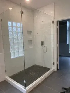 A modern glass-enclosed shower with white tiled walls, a dark grey tiled floor, and a glass block window.