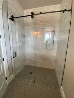 A walk-in shower with a glass sliding door, matte black hardware, and white marble-patterned tile walls and floor.