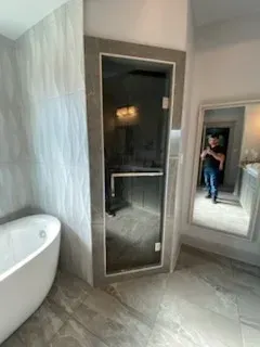 A modern bathroom featuring a white soaking tub, a glass-enclosed sauna, and a mirror reflecting a person.