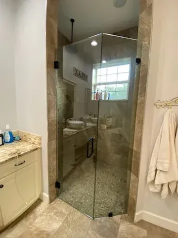 A glass-enclosed, tiled corner shower in a bathroom, situated next to a cream-colored vanity with stone countertop.