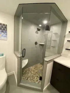 A modern corner shower with a glass door, grey tile walls, a pebble floor, and a small built-in corner seat.