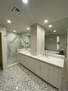 A spacious bathroom featuring a dual vanity with white cabinets, marble countertops, a glass shower, and patterned flooring.