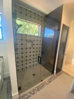 A walk-in shower with grid-patterned tiles, a rectangular window, and a glass door, next to a doorway in a tiled bathroom.