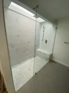 A walk-in shower with white marble-patterned walls, a small built-in bench, glass panel, and a matte black shower head.