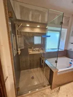 A frameless glass shower enclosure sits next to a bathtub in a bathroom with brown tiled walls and marble flooring.