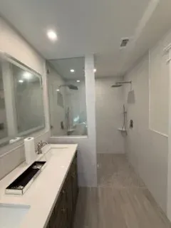 A modern bathroom with a double vanity and mirror on the left, next to a glass-enclosed walk-in shower with light tiles.