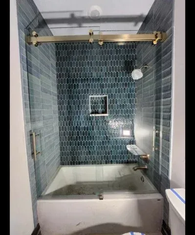 A modern bathtub with blue textured subway tiles, gold fixtures, a sliding glass door, and a built-in shower niche.