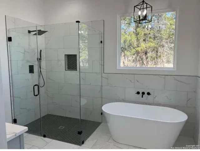 Modern bathroom with a glass-enclosed walk-in shower, a white freestanding soaking tub, and a window overlooking trees.