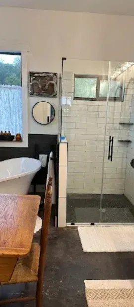 A bright bathroom with a freestanding white tub, wooden furniture, and a glass-enclosed shower with white subway tiles.