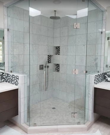 A modern corner shower with glass walls, marble-style tile, an overhead rainfall showerhead, and small recessed shelving.