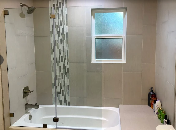 Modern bathroom with a white bathtub, glass shower screen, neutral tiled walls with a vertical mosaic accent, and window.