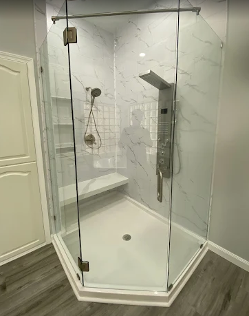 A corner shower stall with a white base, marble-patterned walls, a glass enclosure, a built-in bench, and a panel shower.
